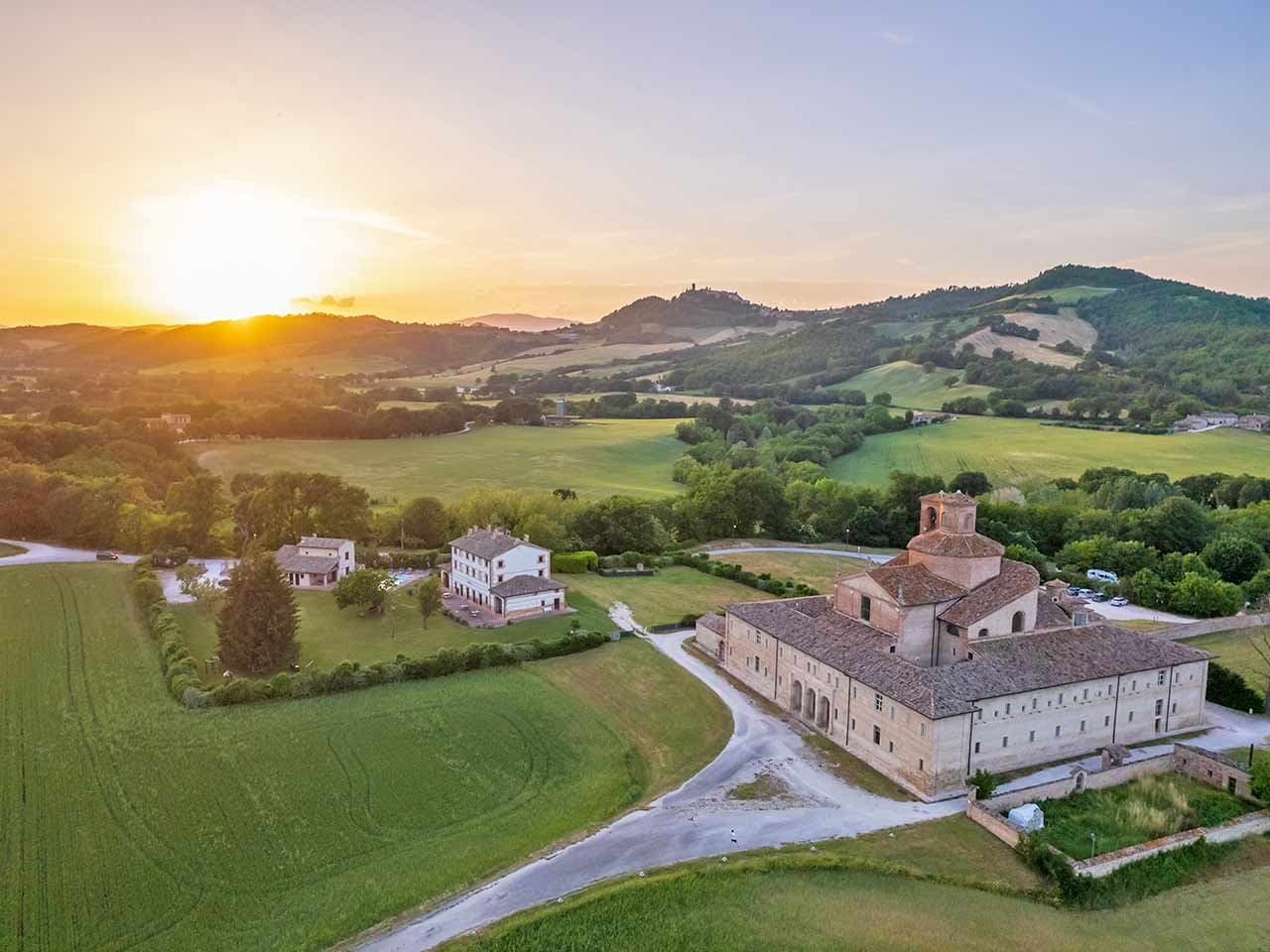 Veduta aerea del Montefeltro al tramonto, con un palazzo storico e una villa immersi nella campagna collinare