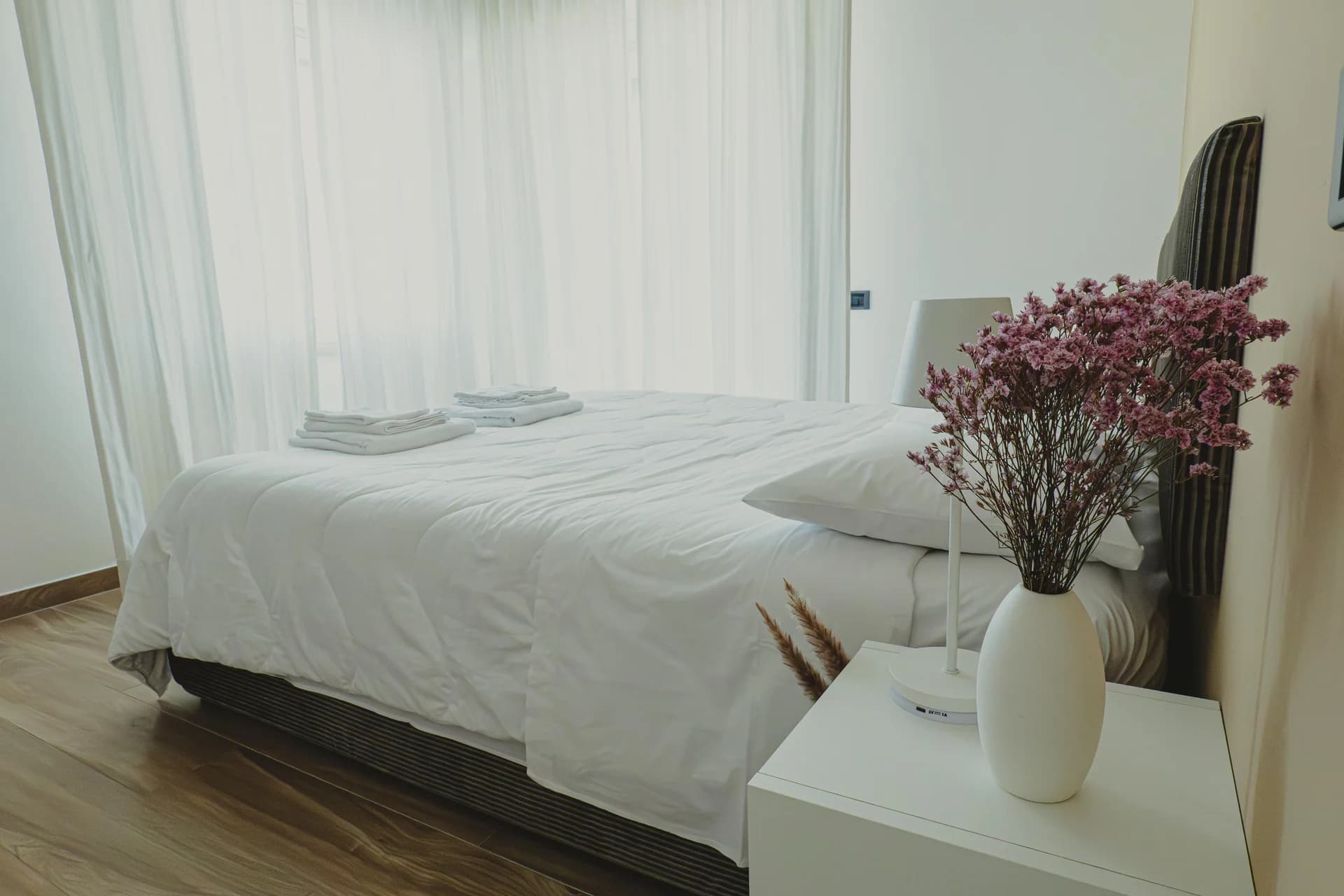 Bedroom dressed in white in natural light, with a vase of pink flowers on the bedside table and sheer white curtains