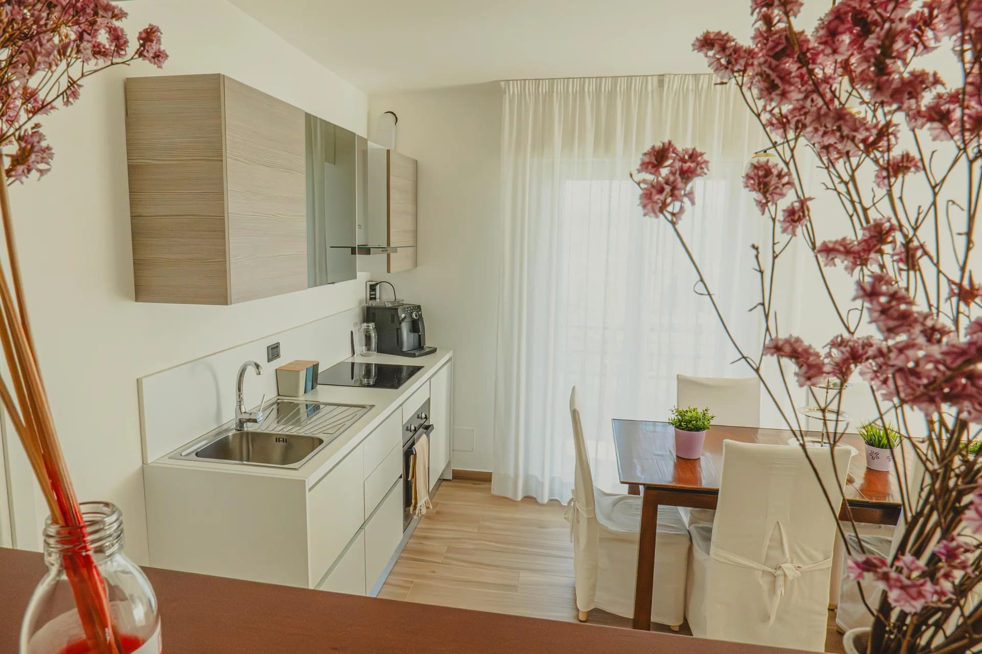 Cucina moderna bianca con piano cottura, macchina da caffè e tavolo da pranzo in legno accanto alla finestra con tende chiare