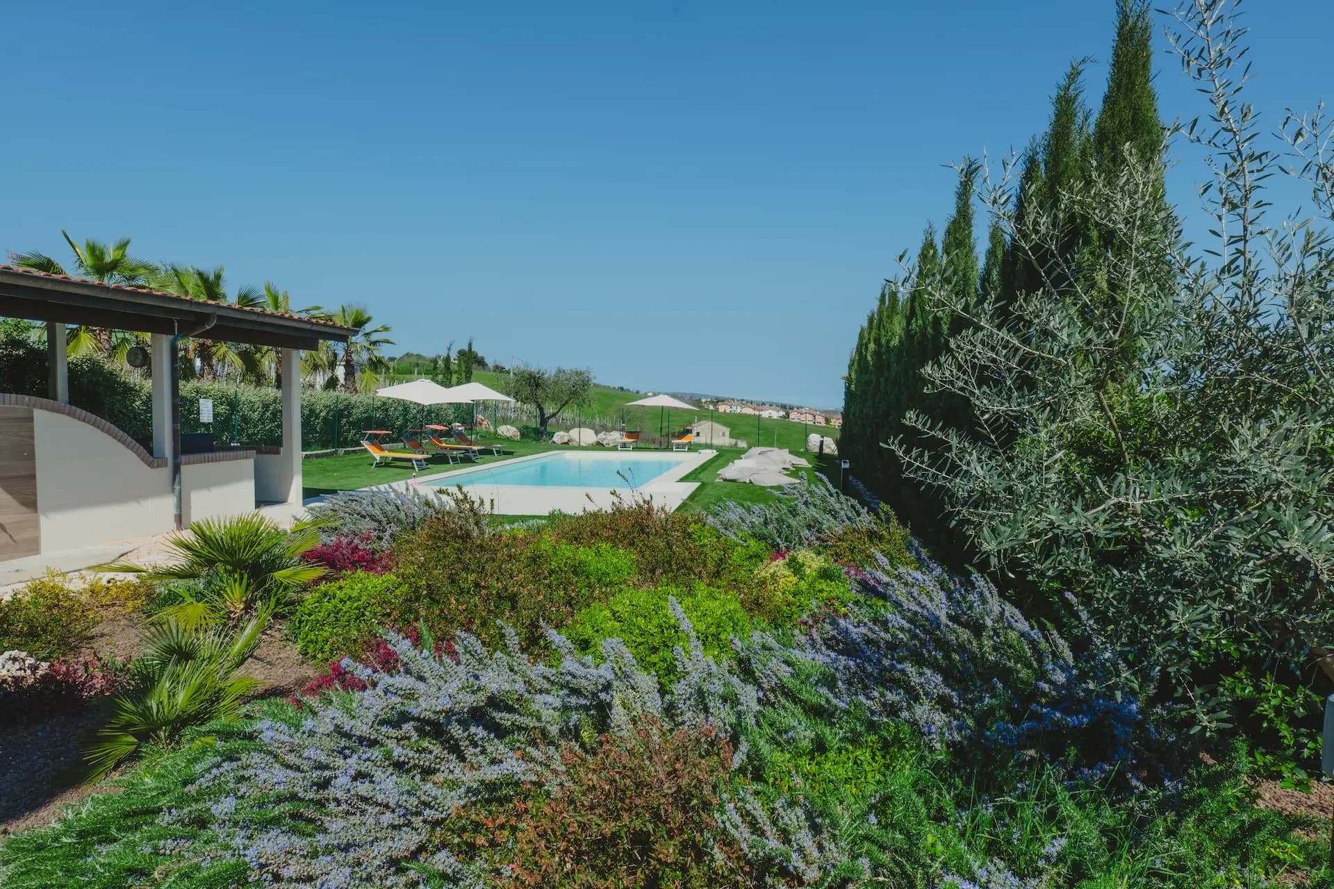 La piscina vista attraverso un giardino fiorito di lavanda e rosmarino, con ulivi, cipressi e colline sullo sfondo