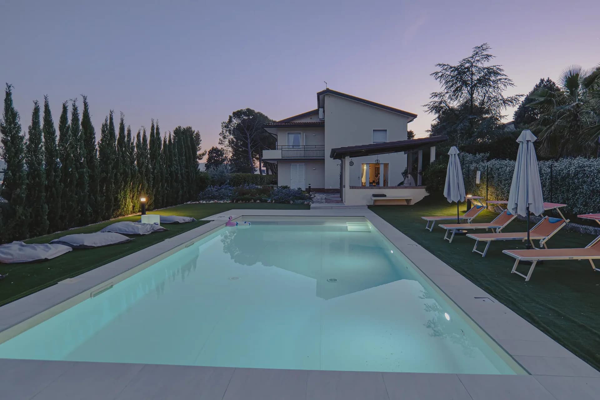 La piscina illuminata al crepuscolo davanti alla villa, con cielo viola, lettini e ombrelloni chiusi