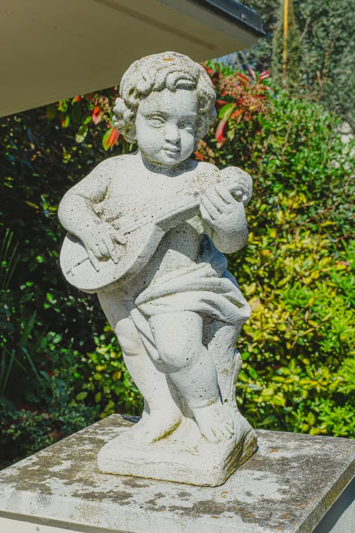 Statua in pietra di un cherubino che suona un mandolino, posata su un piedistallo tra il verde del giardino