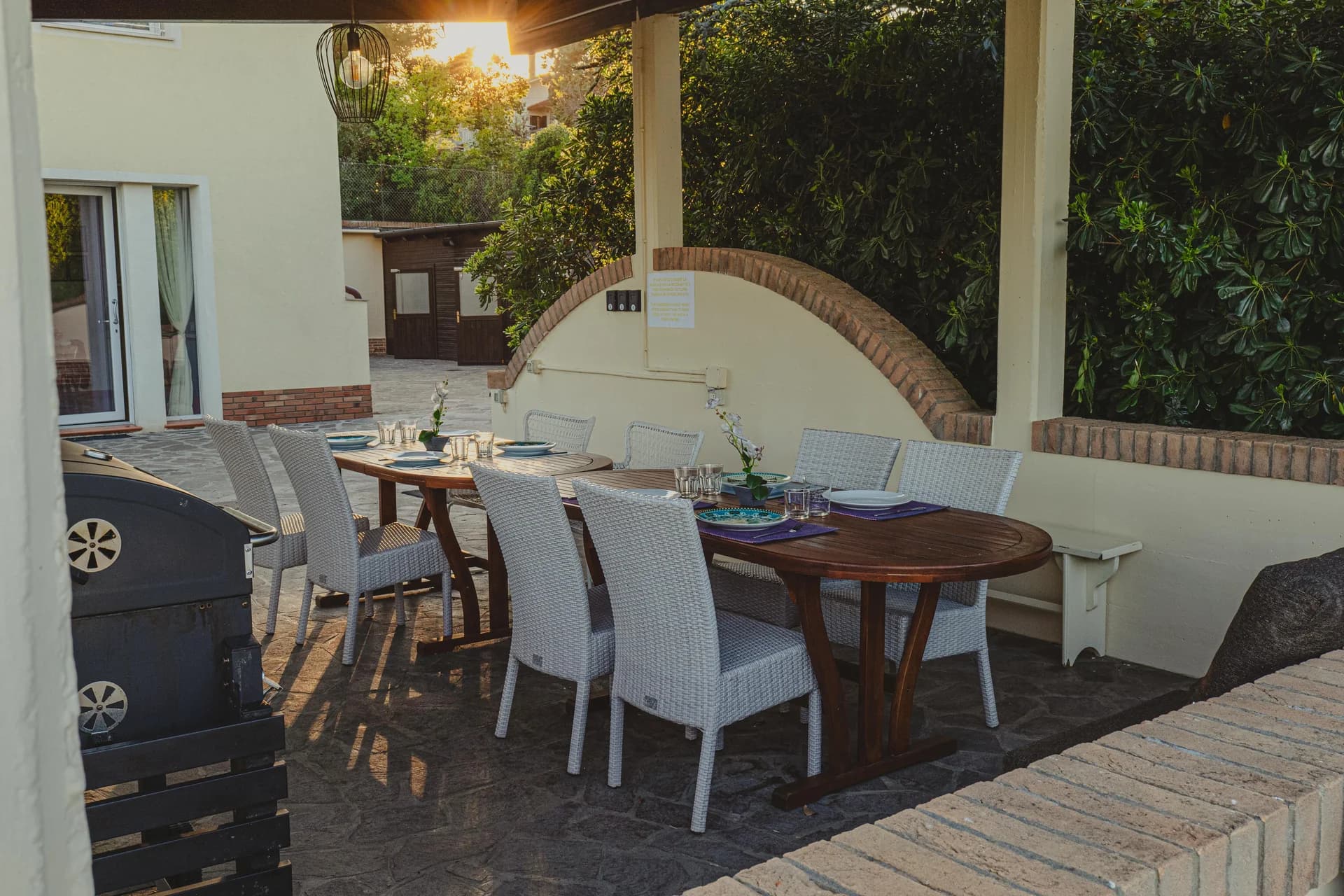 Terrazza esterna coperta con due tavoli apparecchiati, sedie in rattan chiaro e barbecue, al tramonto