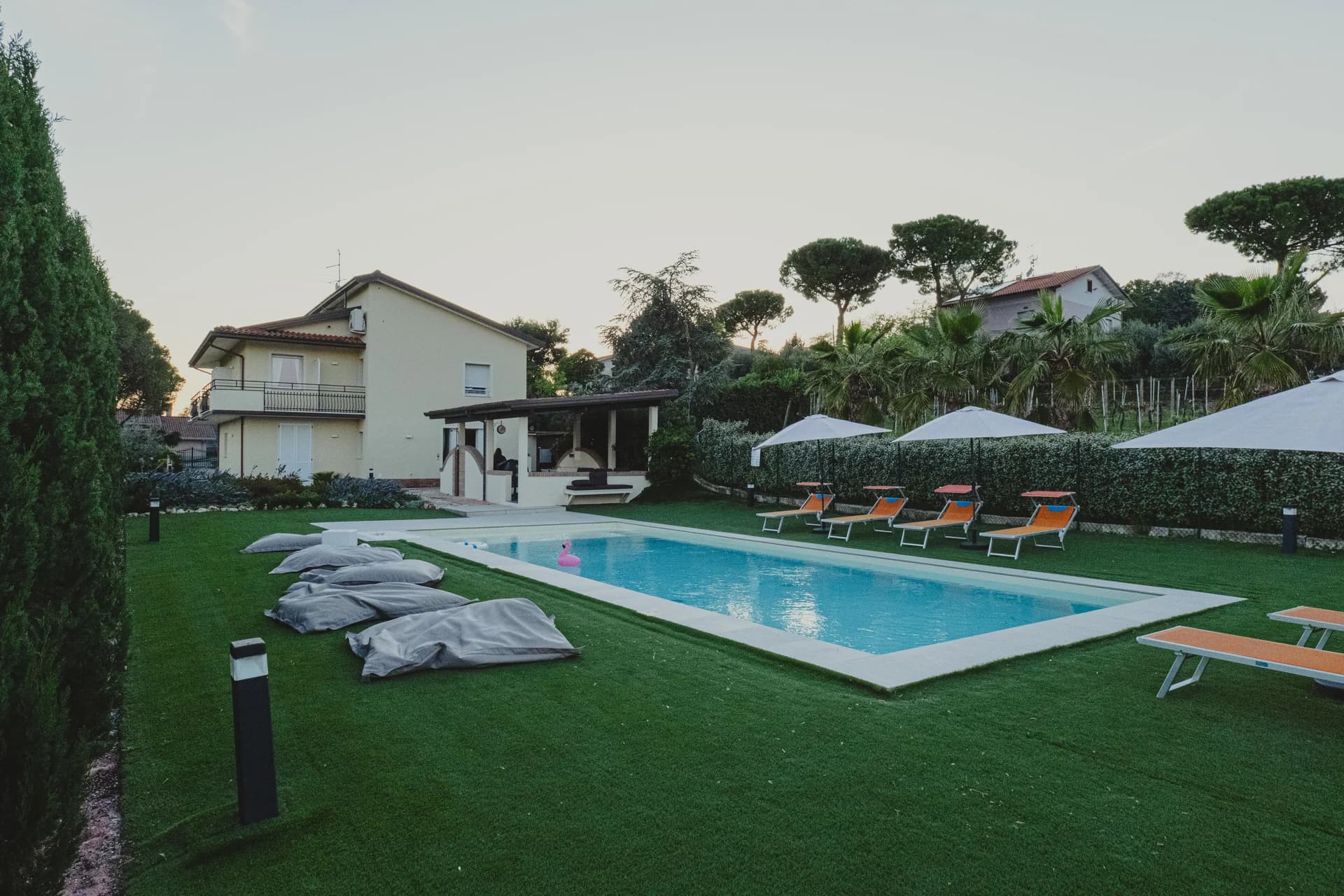 The villa and pool in the garden at dusk, with orange loungers, bean bags on the lawn and palm trees